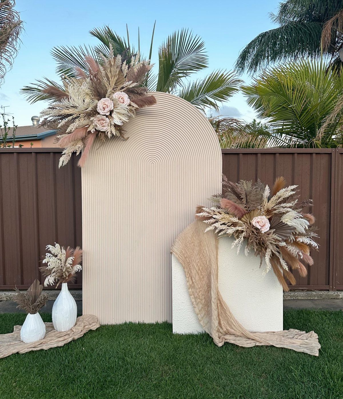 Beige arched wedding backdrop decorated with pampas grass and pink roses outdoors.