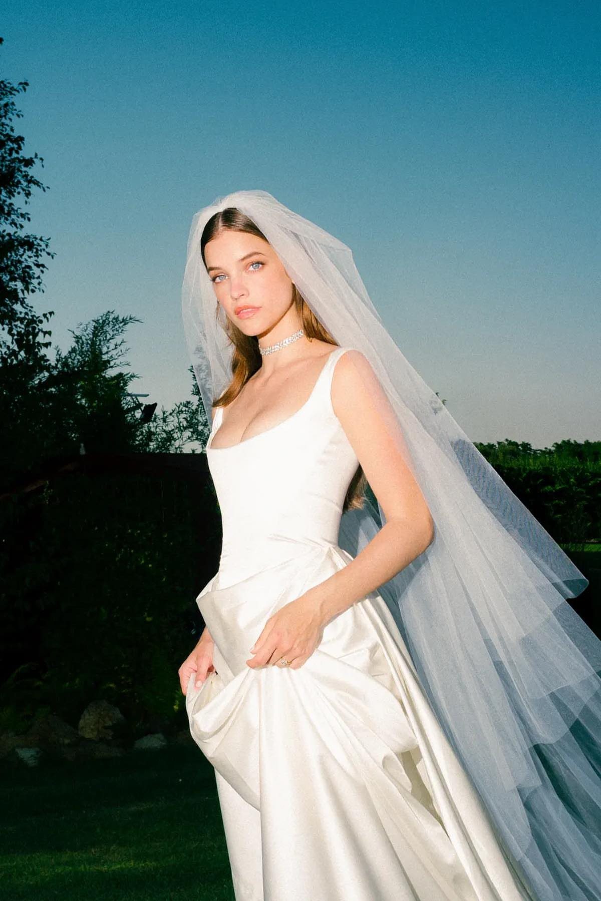 Barbara Palvin in a white satin wedding dress and long tulle veil outdoors.