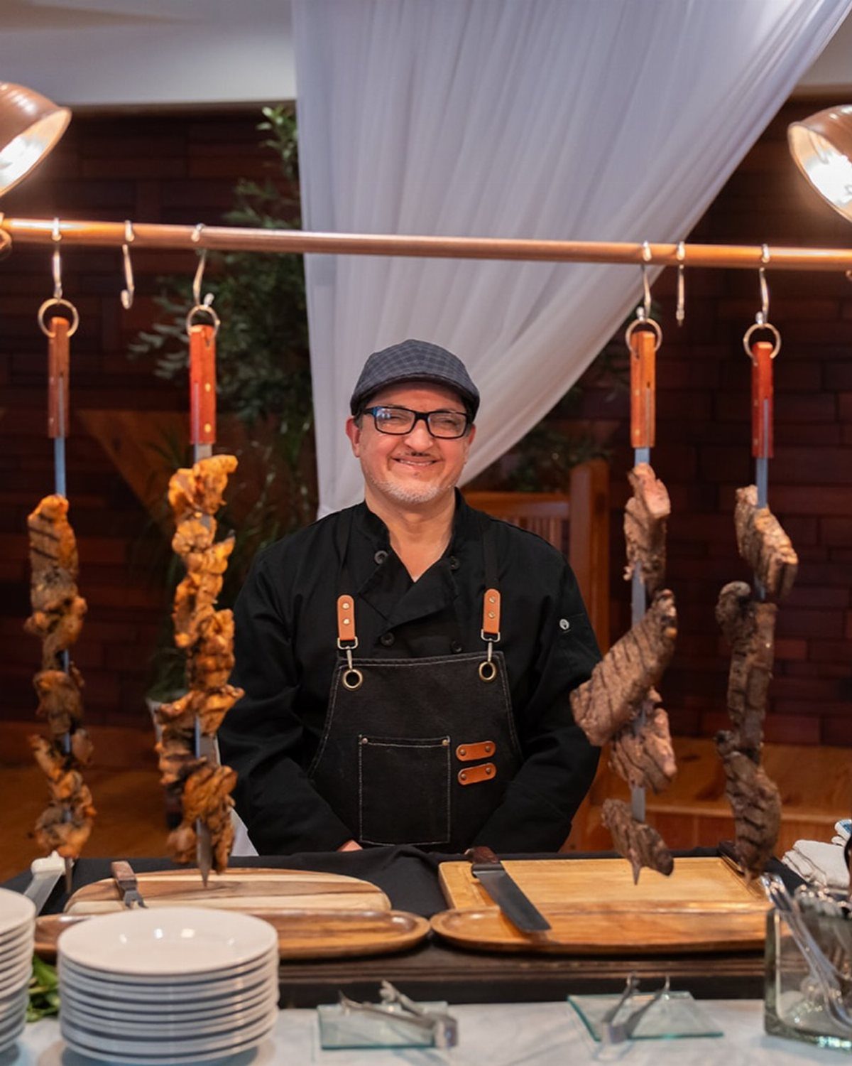 Chef standing behind a grilling station with hanging meats and wooden serving boards.