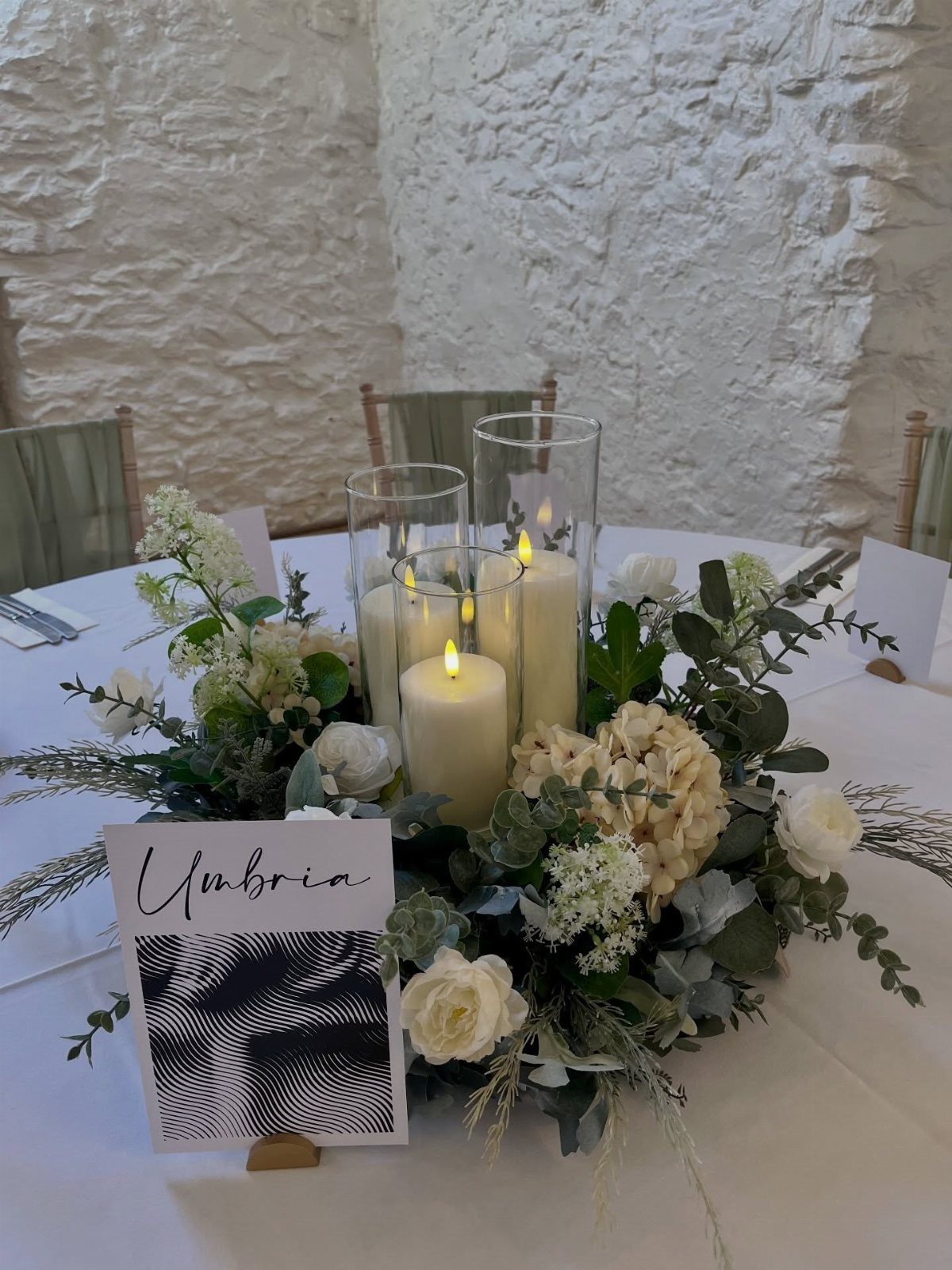 White pillar candles in glass cylinders surrounded by white roses and greenery on a table.