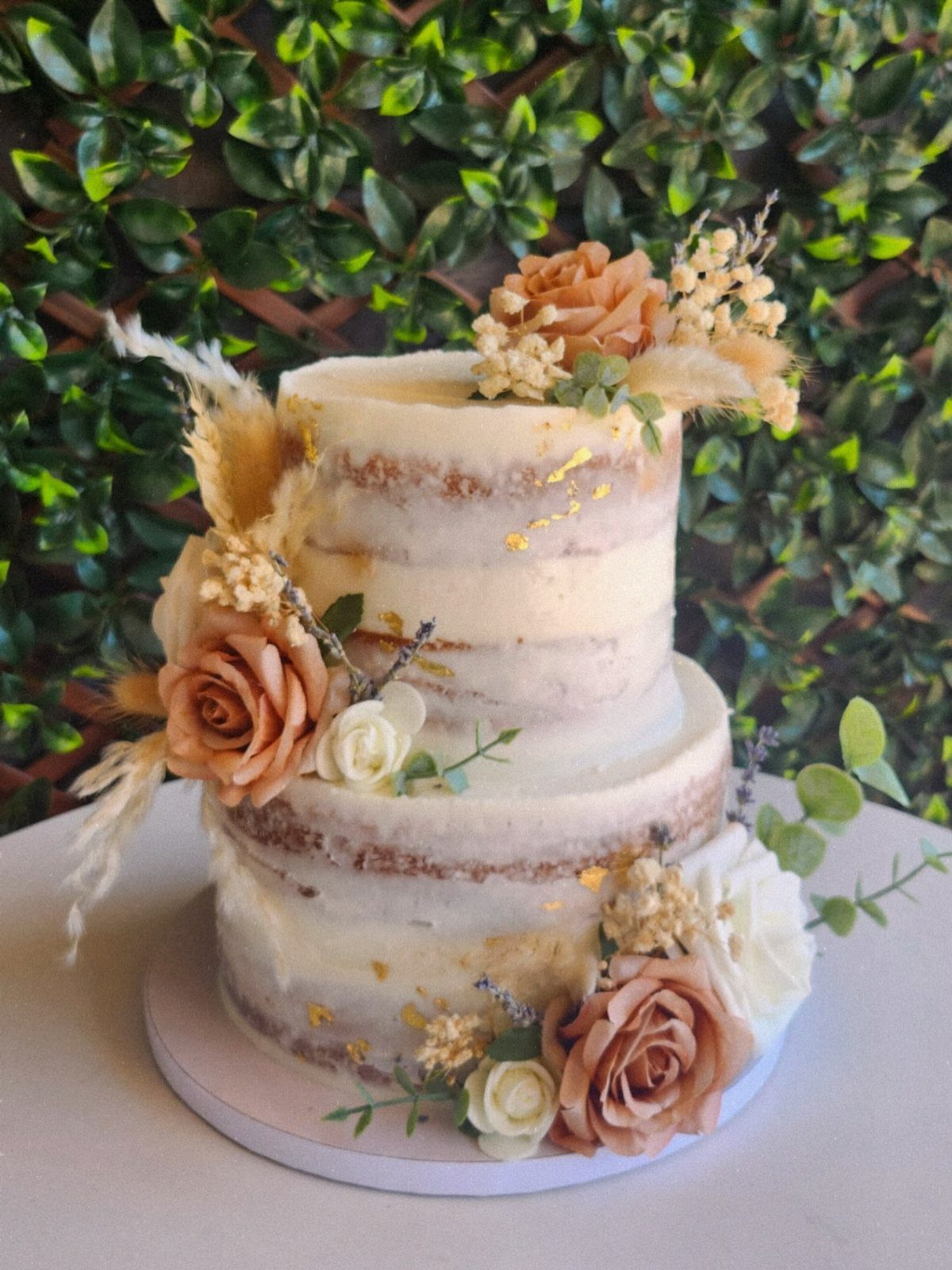 Semi-naked two-tier wedding cake decorated with peach roses and gold leaf against greenery.