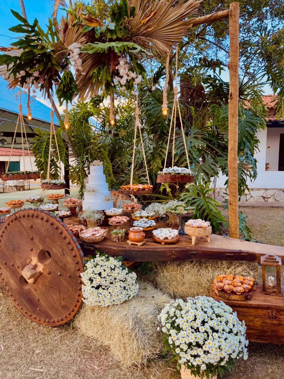 Rustic outdoor dessert table with sweets, white cakes, and a large wooden wagon wheel.