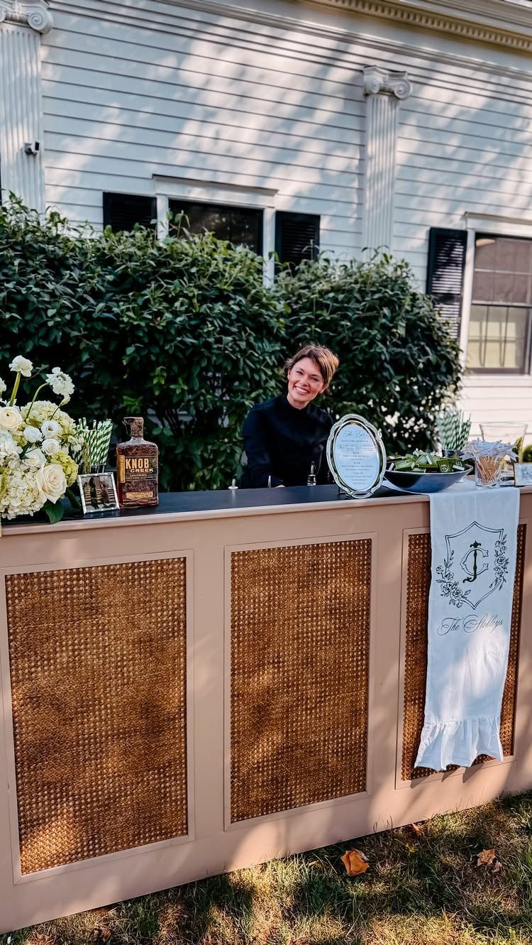 Woman smiling behind a tan outdoor bar with woven cane panels and white flowers.