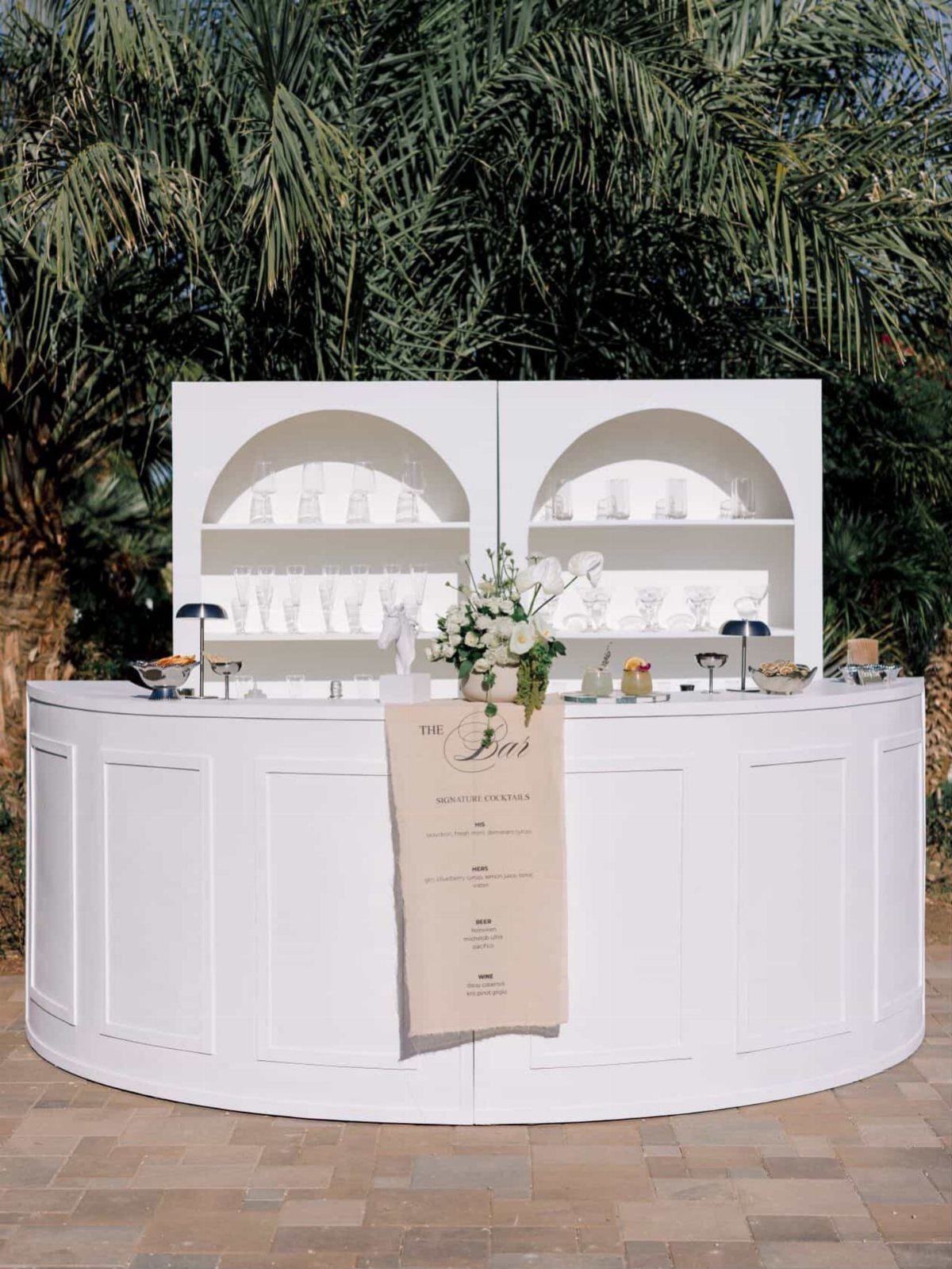 White curved outdoor wedding bar with glassware and floral arrangements against palm trees