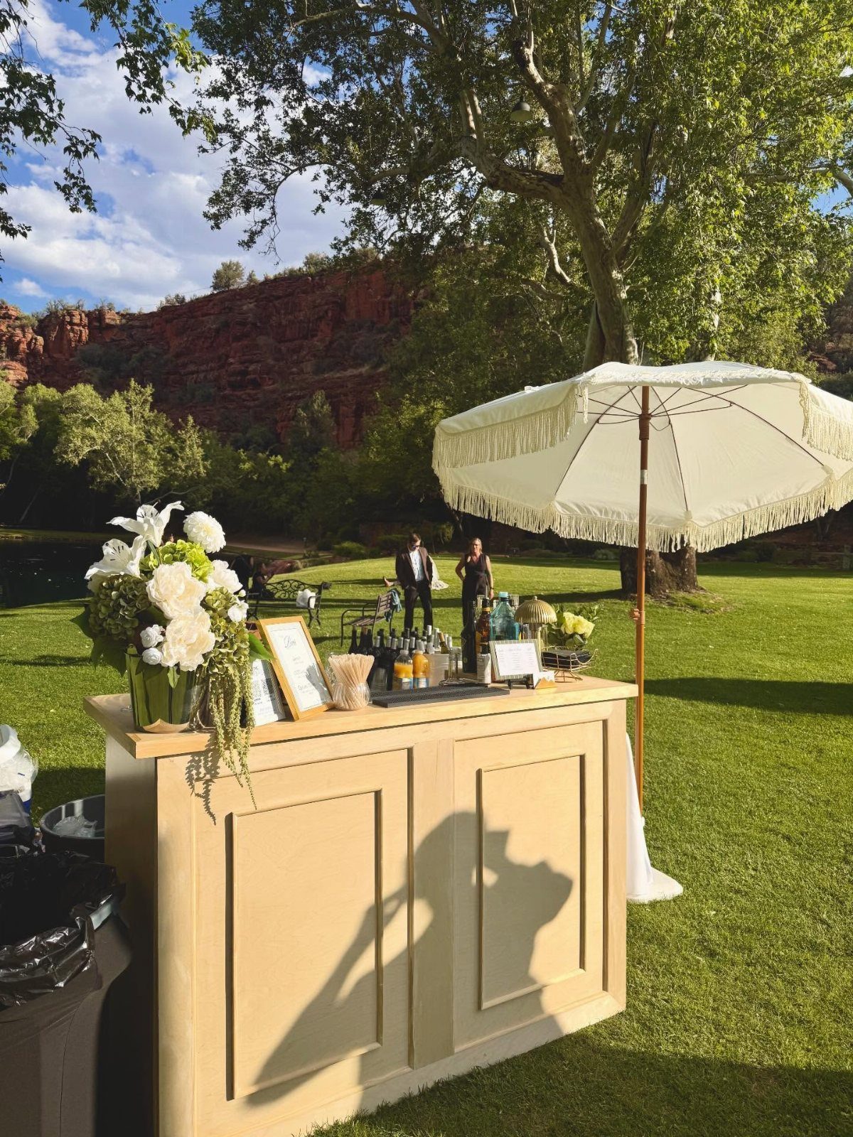 Outdoor wedding bar with cream counter, white flowers, and a fringed umbrella.