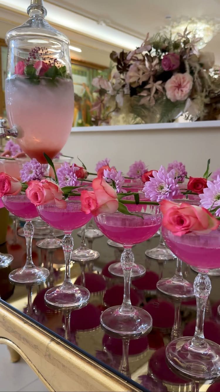 Pink drinks in coupe glasses with rose garnishes on a mirrored gold table.