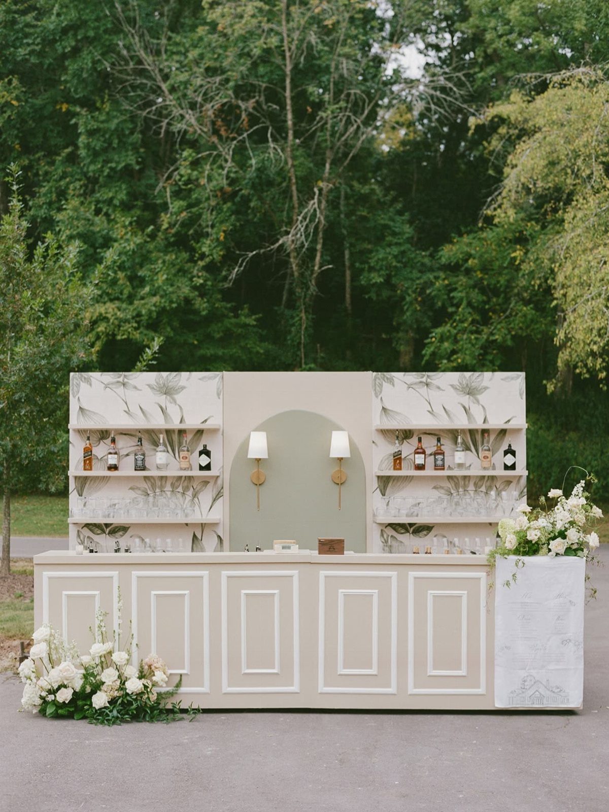 Cream-colored outdoor wedding bar with liquor bottles on botanical printed shelves.