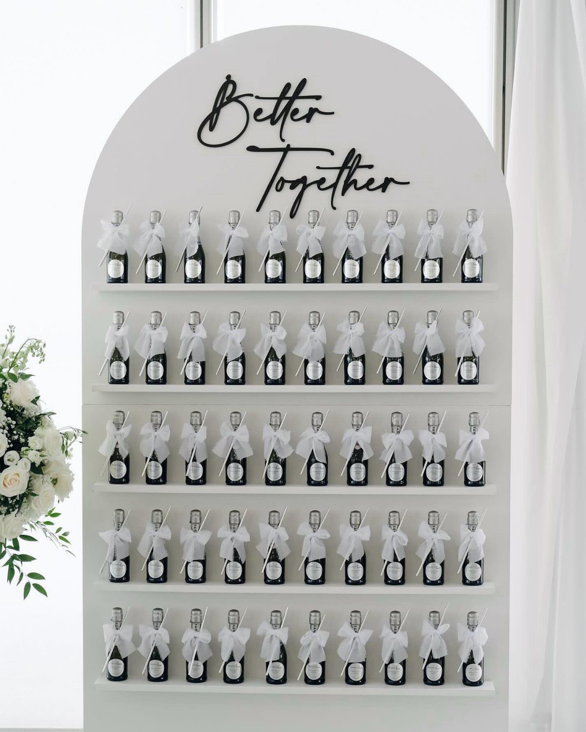 Champagne bottles with white bows on a white arched wall saying Better Together