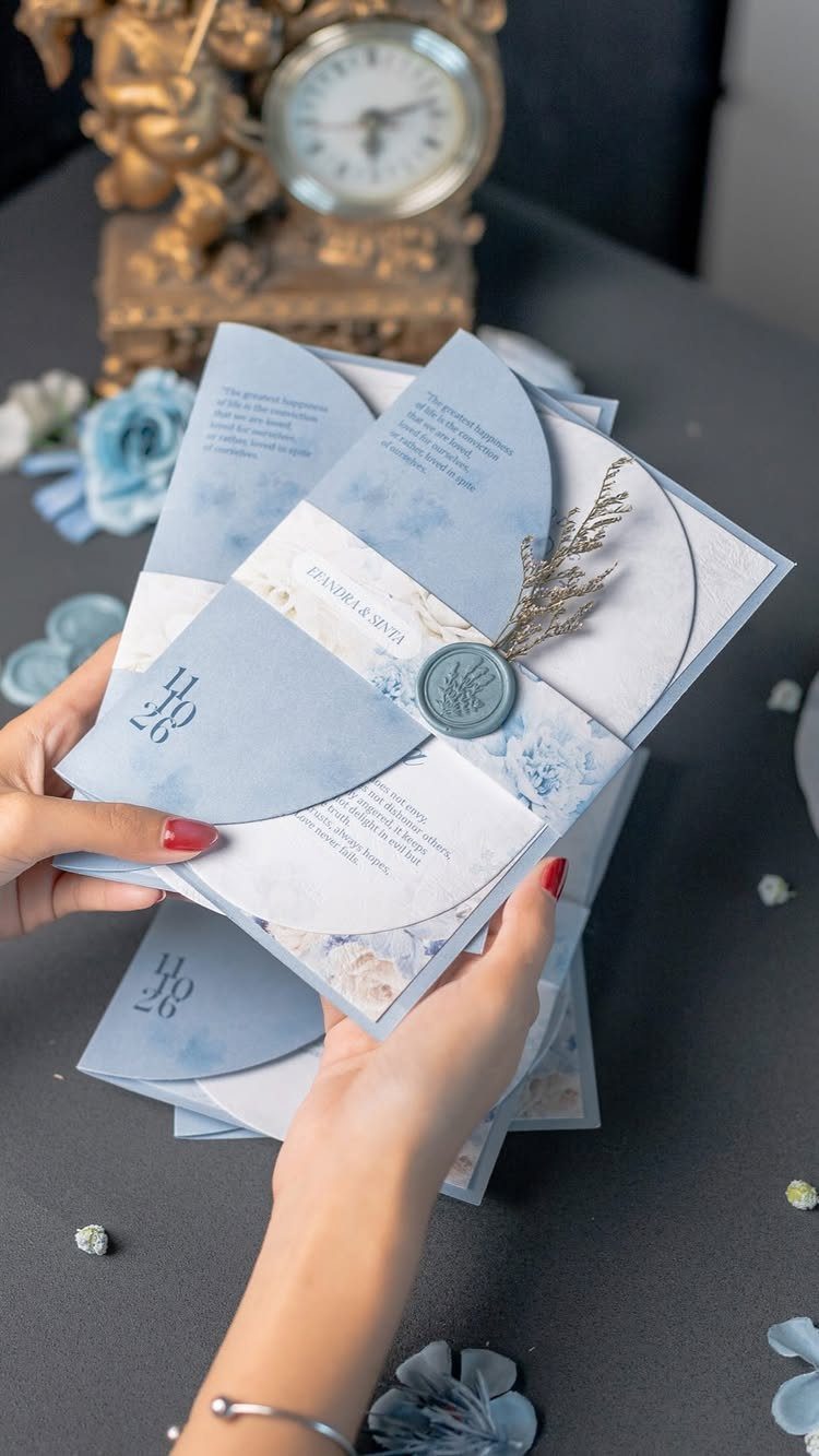 Dusty blue and white wedding invitations with a wax seal held by hands.
