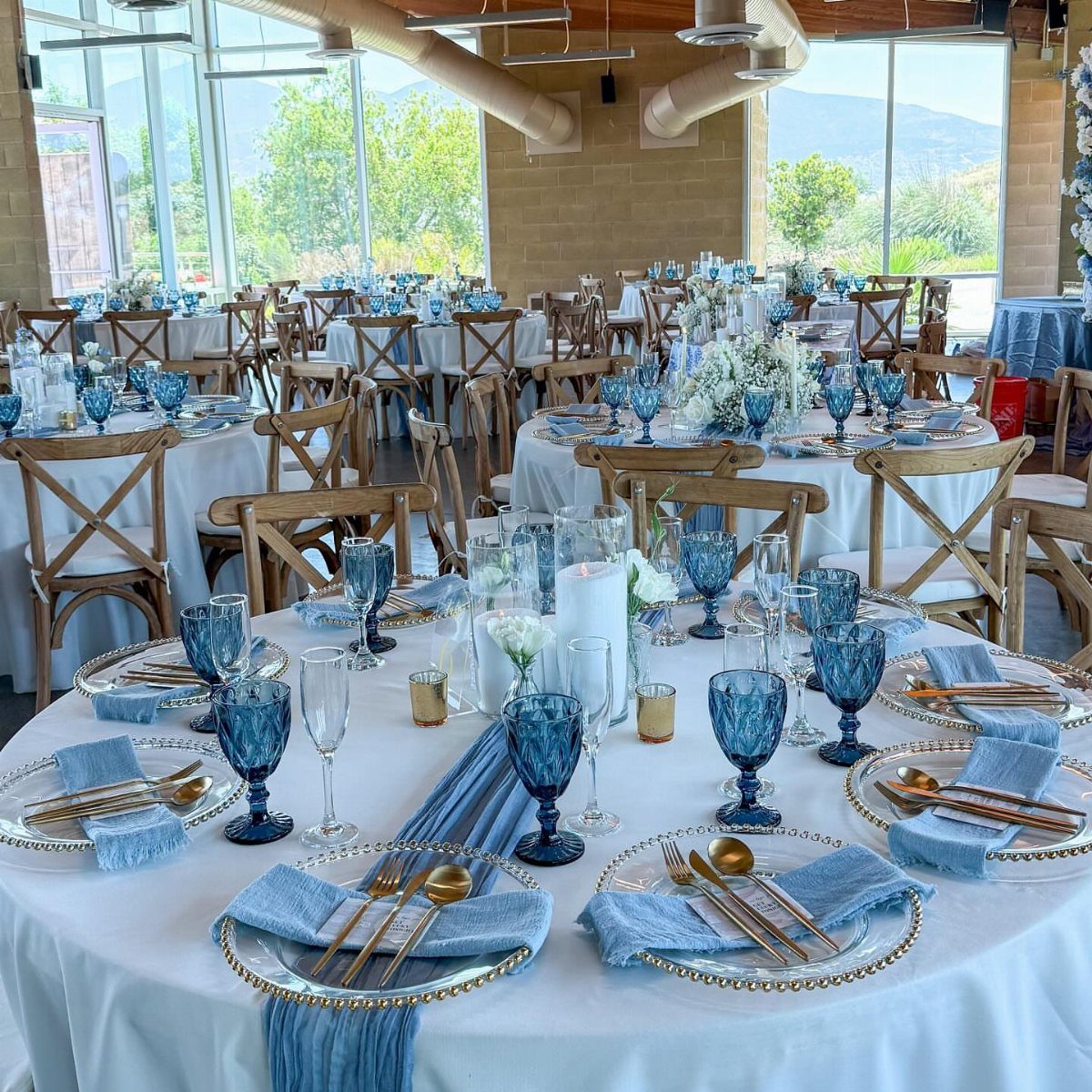 Wedding reception tables with white linens, dusty blue runners, and gold cutlery.