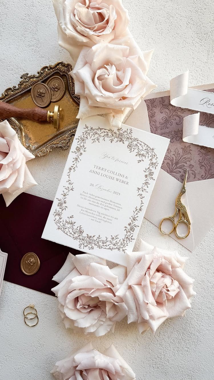 Blush roses and a burgundy envelope surrounding a white wedding invitation with gold accents.