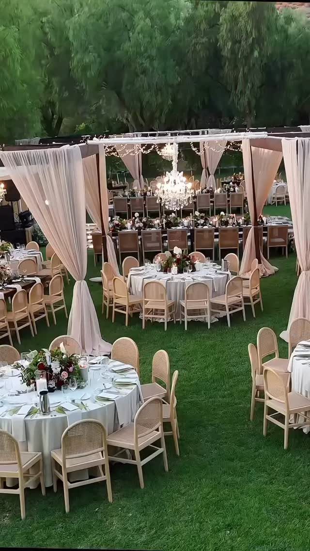 Outdoor wedding reception with blush fabric draping, white tables, and light wood chairs.