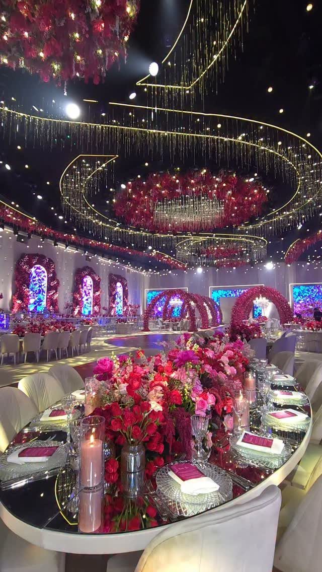 Lavish wedding reception hall with red and pink floral centerpieces and gold ceiling lights.