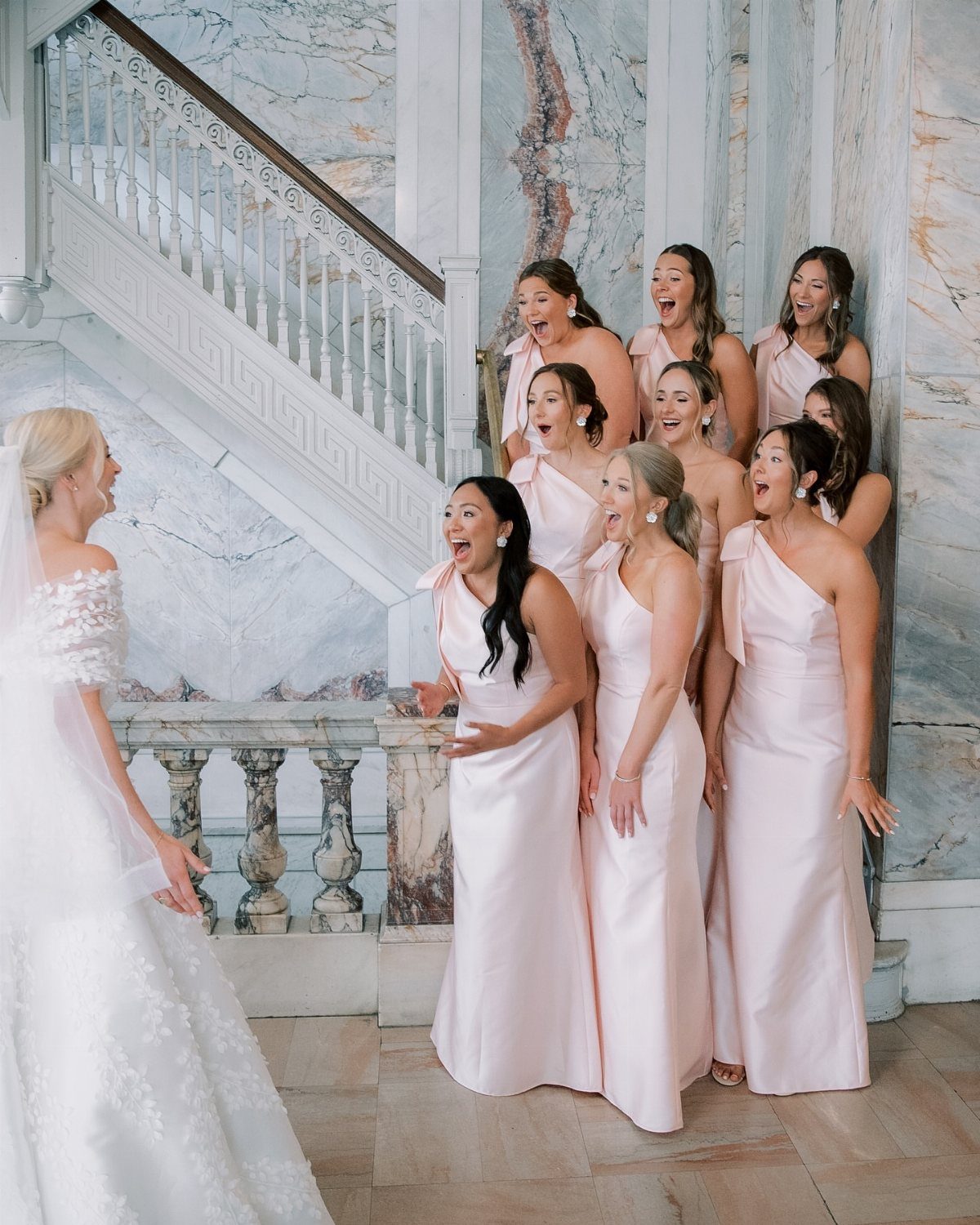 Bridesmaids in blush pink satin gowns reacting to a bride in white lace.