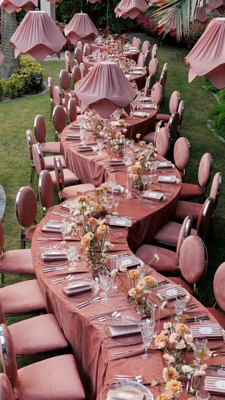 Winding outdoor banquet table with blush pink linens, chairs, and hanging fabric lampshades.