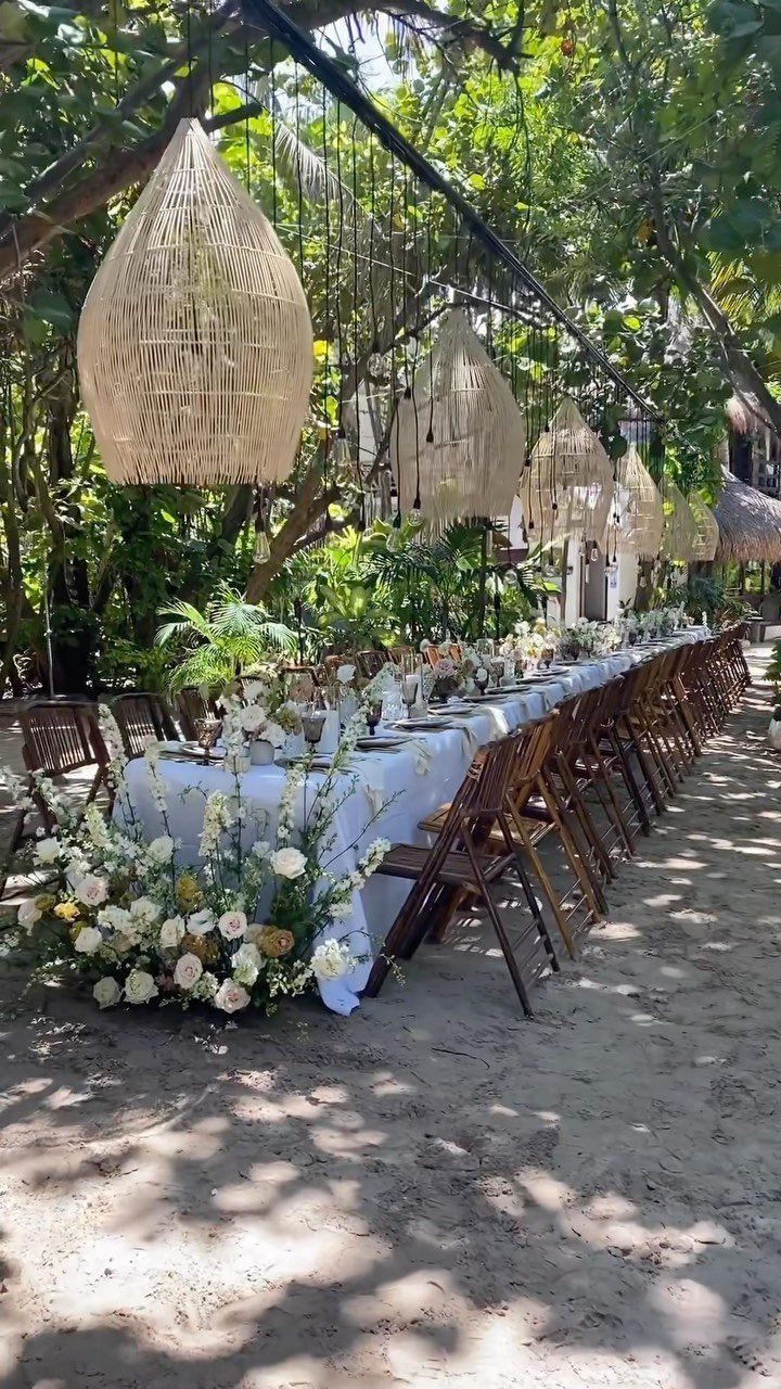 Woven wicker lanterns hanging over a long white wedding table on a beach