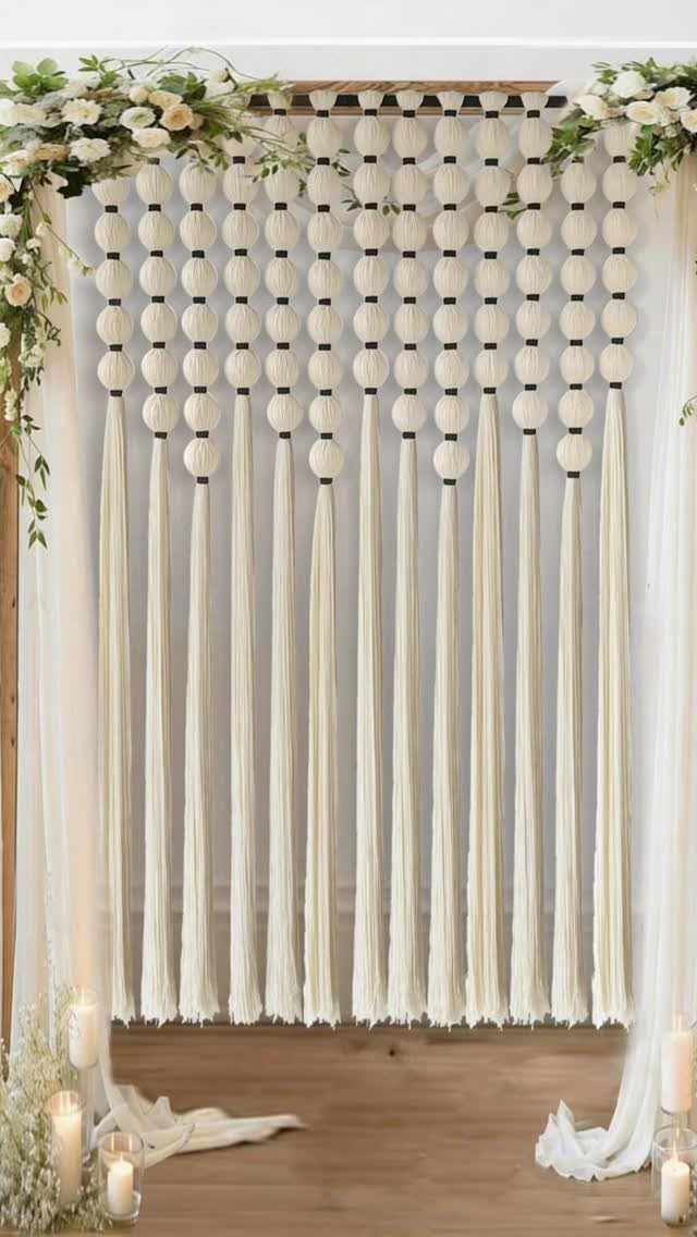 Cream macramé backdrop with knotted spheres and tassels framed by white floral greenery.