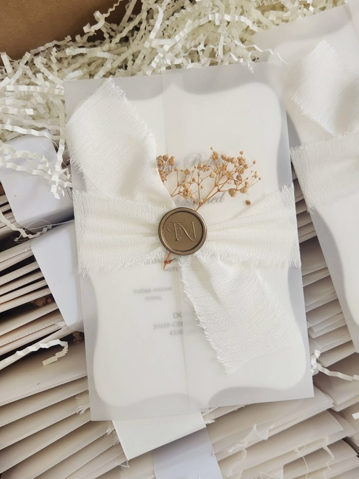 White wedding invitation with a sheer ribbon, bronze wax seal, and dried flowers.
