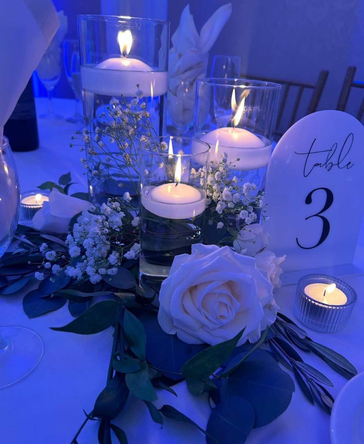 Floating white candles in glass cylinders with white roses and greenery on wedding table.