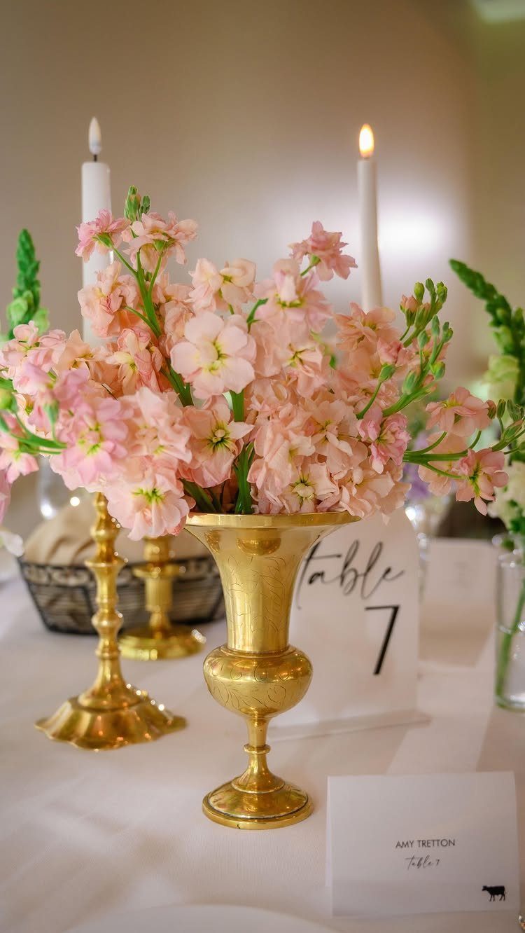 Peach flowers in a gold pedestal vase with lit white taper candles.