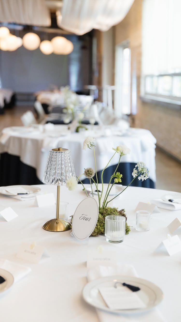 Minimalist wedding centerpiece with white flowers and a gold lamp on a white table.