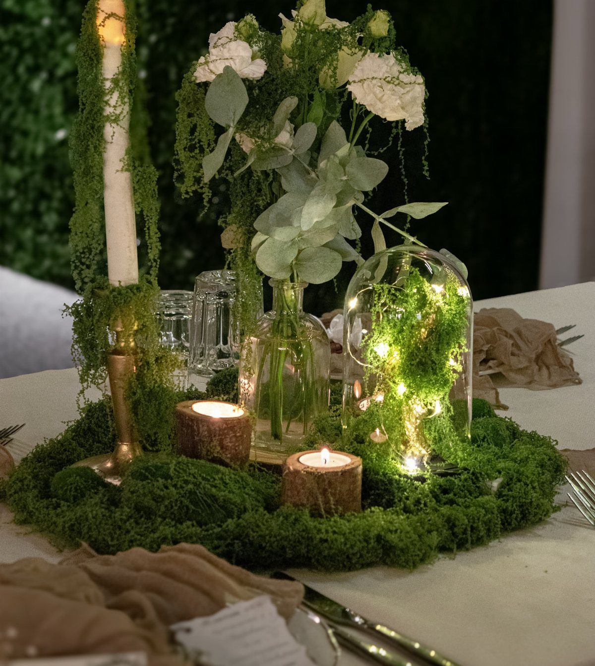 Green moss table runner with white flowers, lit candles, and fairy lights in glass.