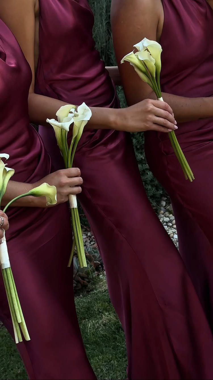 Bridesmaids in burgundy satin dresses holding white calla lily bouquets outdoors.