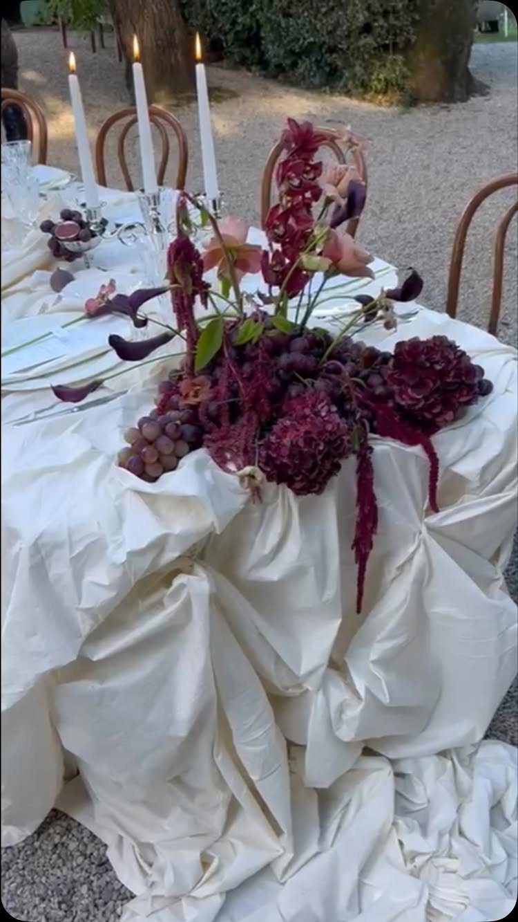 Burgundy floral centerpiece with purple grapes on a draped white tablecloth with candles.