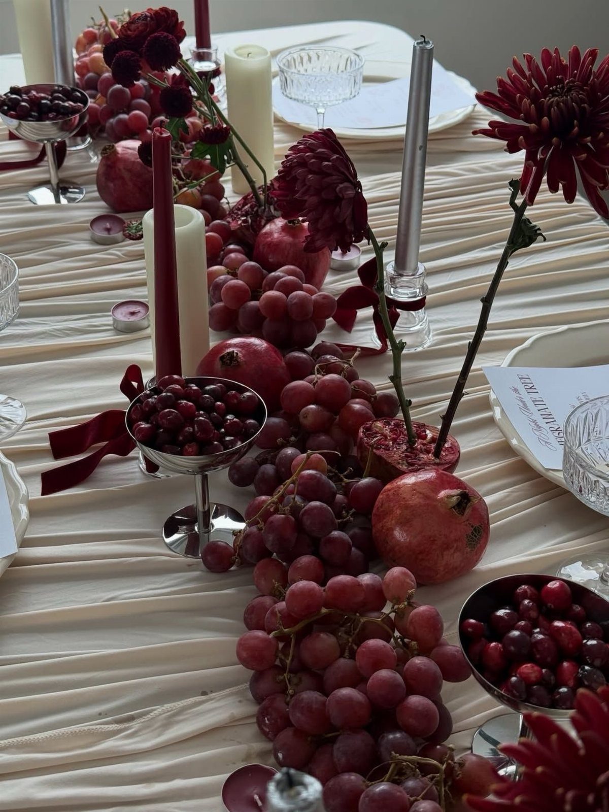 Burgundy grapes, pomegranates, and red flowers arranged as a table runner with candles.
