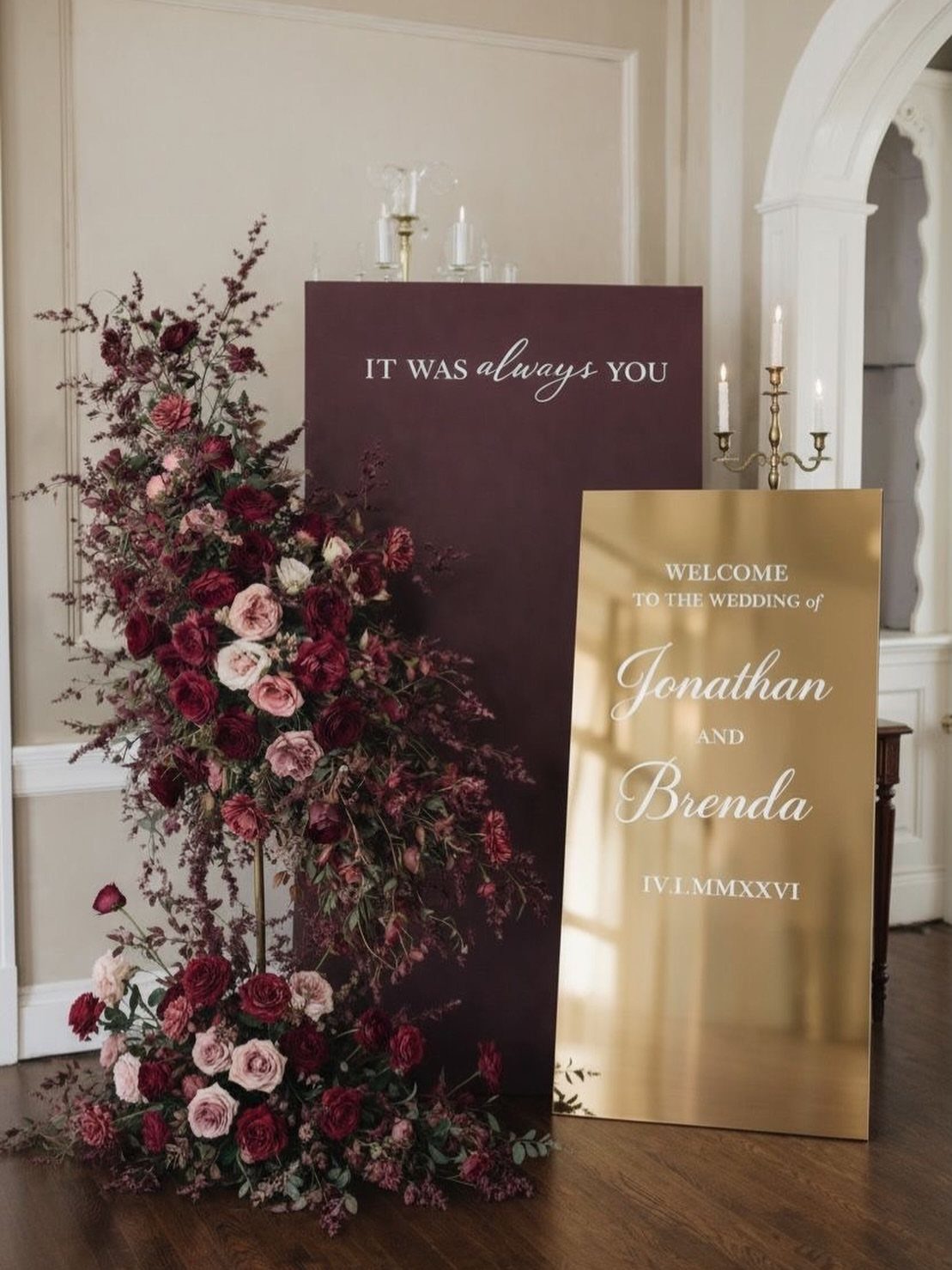 Burgundy and pink floral arrangements framing a gold mirrored wedding welcome sign.