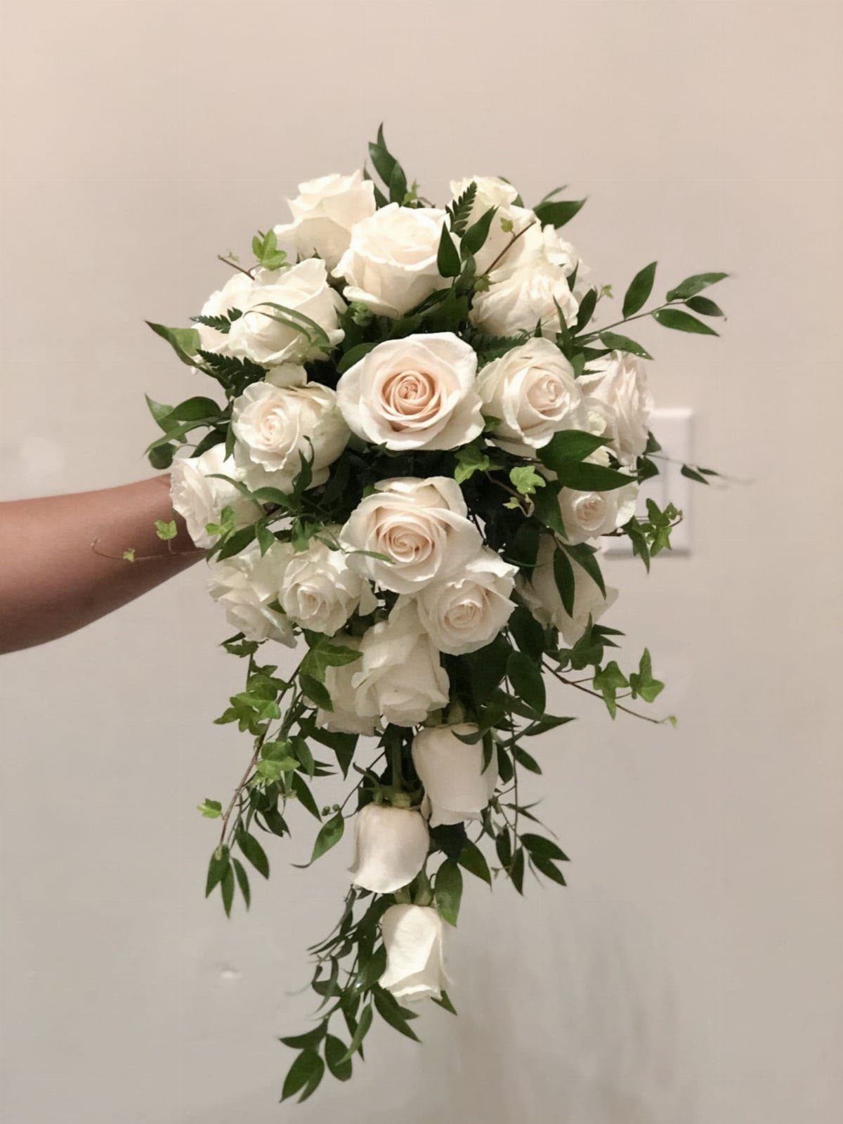 Cascading bridal bouquet of white roses and green leaves held against a wall