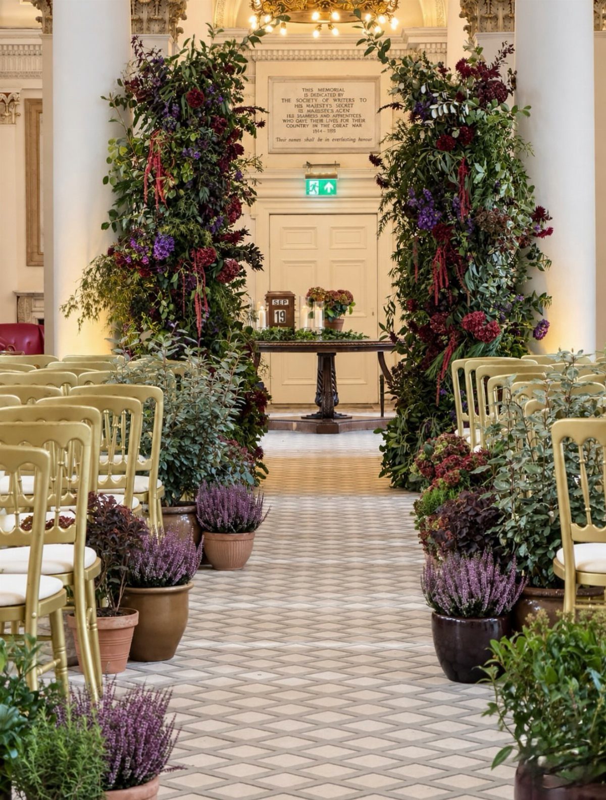 Wedding ceremony aisle with gold chairs and tall dark floral pillars and greenery