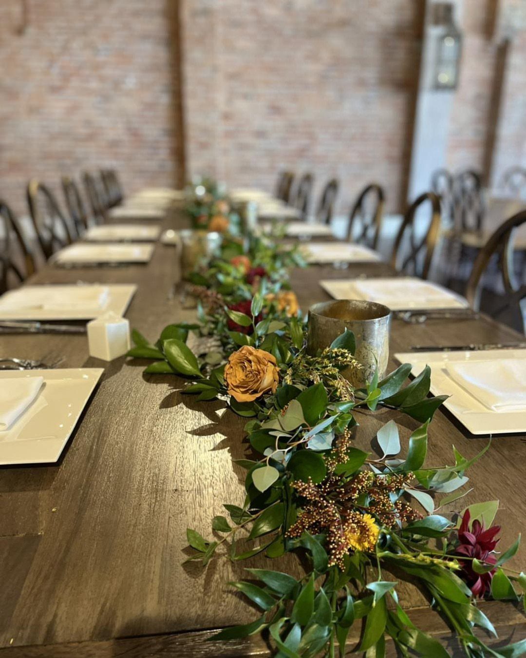 Wedding table runner made of dark greenery and orange flowers on a wooden table.