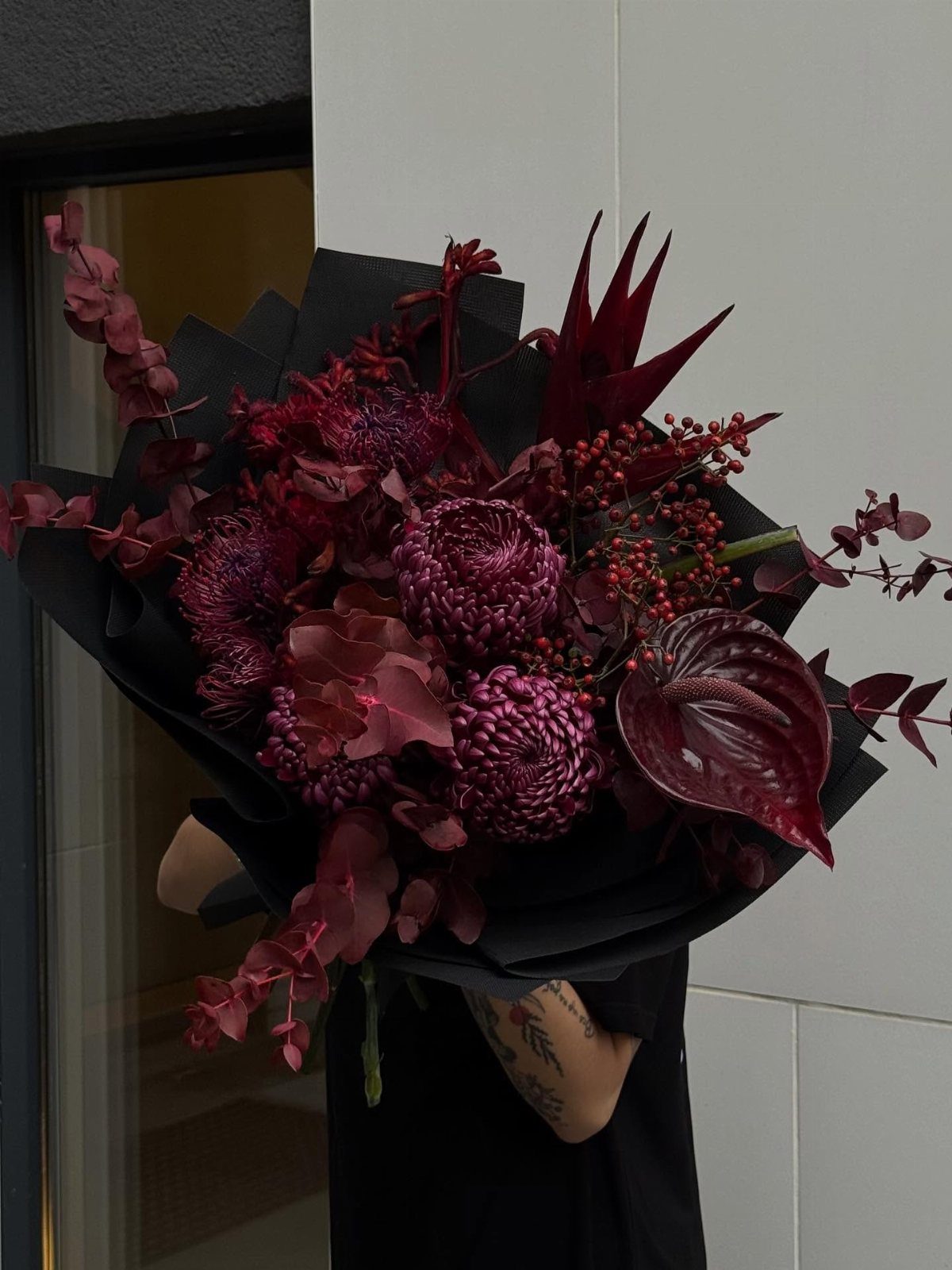 Deep burgundy and maroon floral bouquet wrapped in black paper held by person