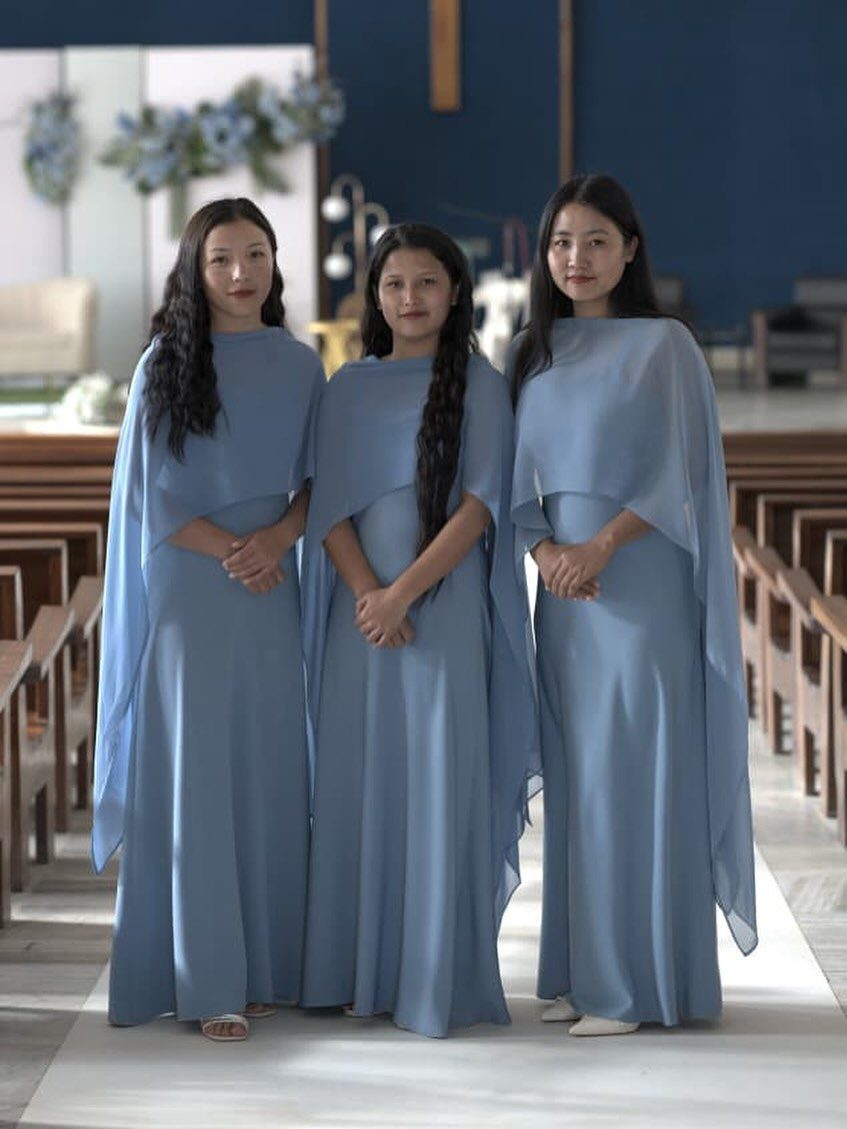 Three women wearing matching dusty blue bridesmaid dresses with capes in a church.