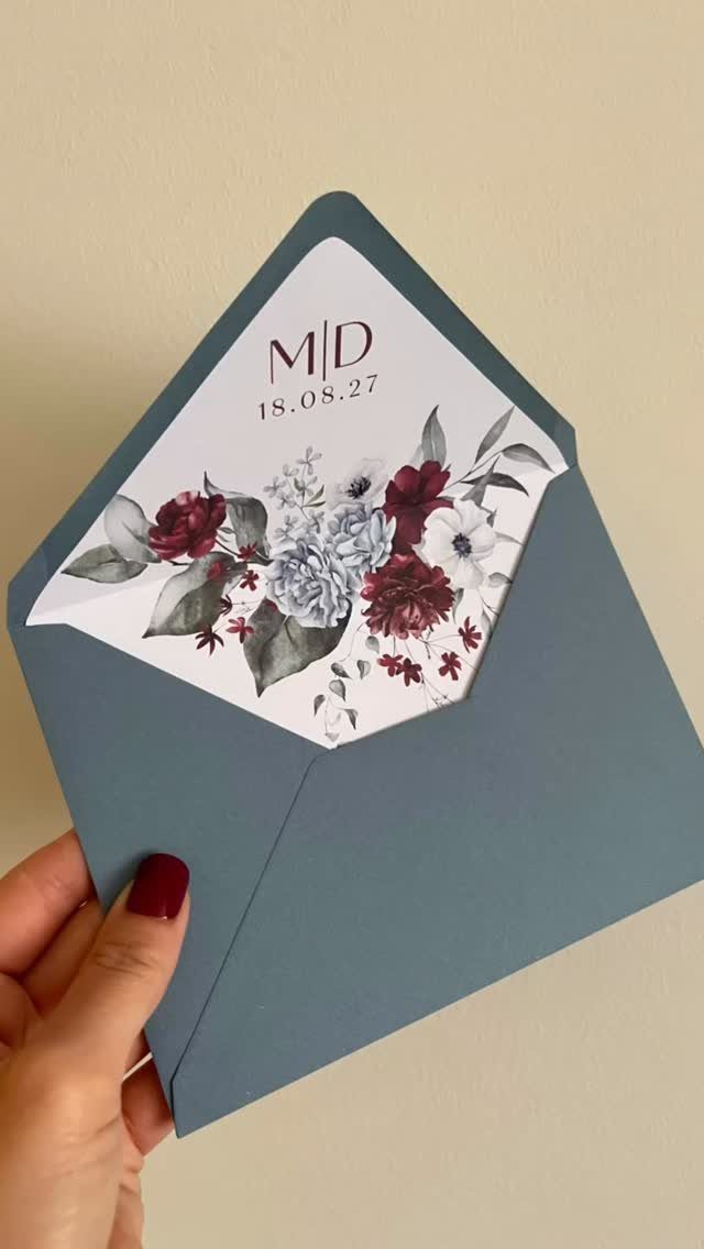 Dusty blue envelope holding a white wedding invitation with burgundy floral designs.