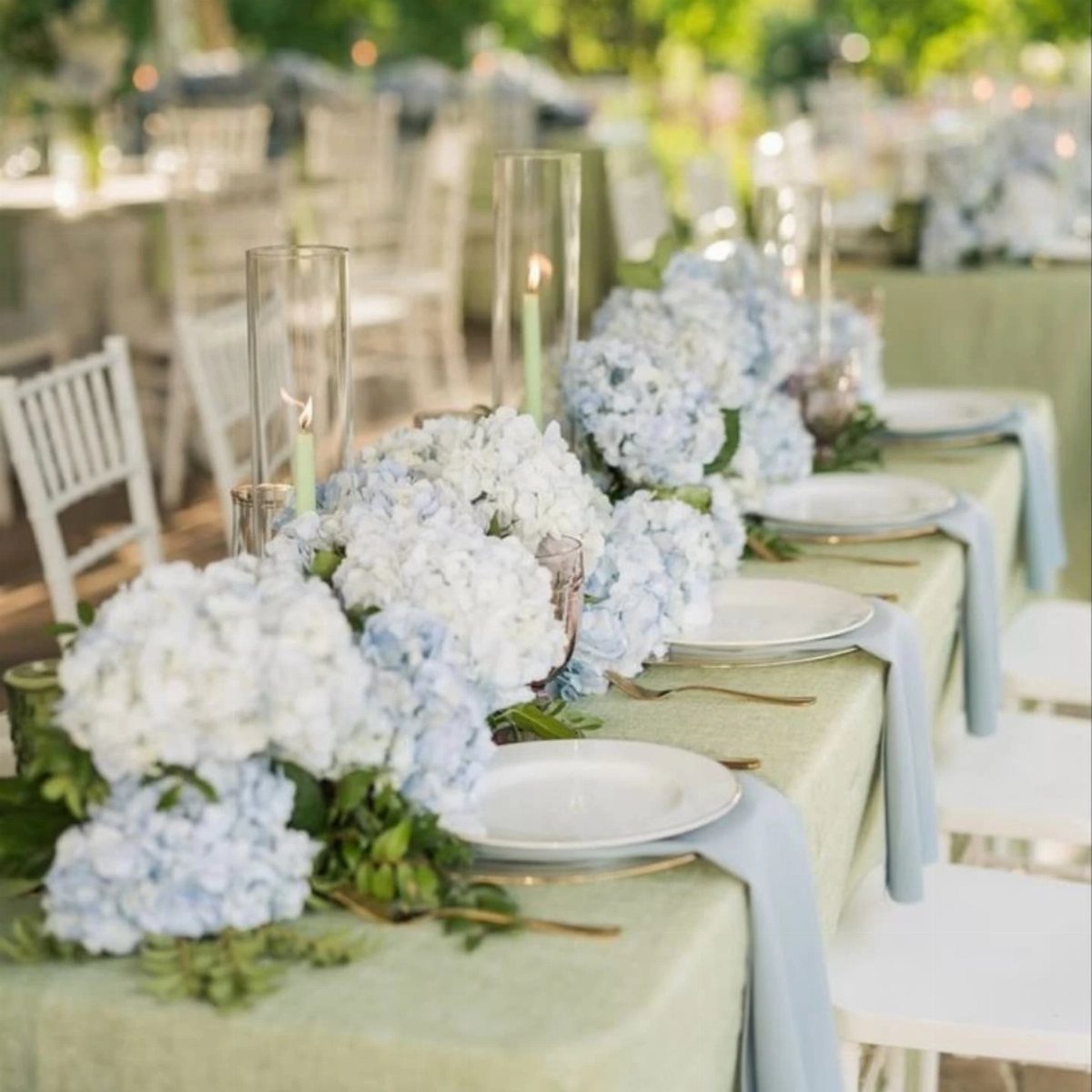 Sage green table with dusty blue hydrangea floral runner and blue napkins outdoors.