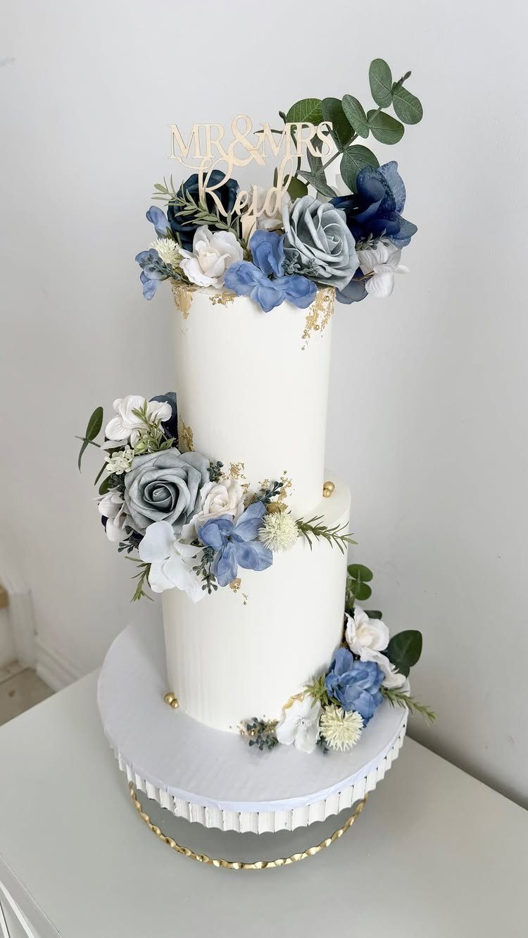 Two-tier white wedding cake decorated with dusty blue roses and gold topper.