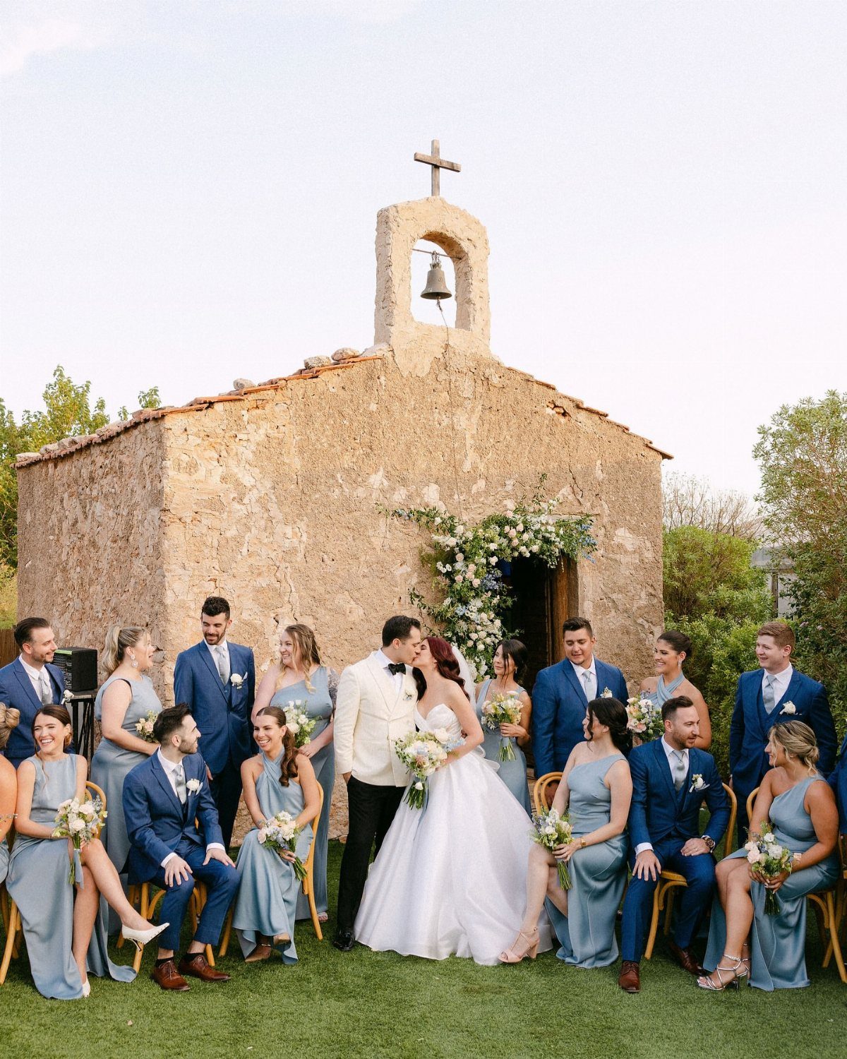 Wedding party in dusty blue attire posing before a rustic stone chapel.