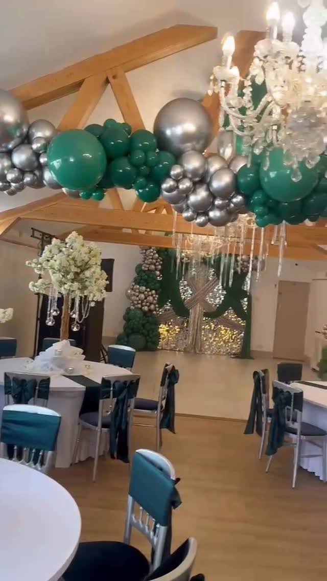 Event room with emerald green and silver balloons, white tables, and green chair sashes.