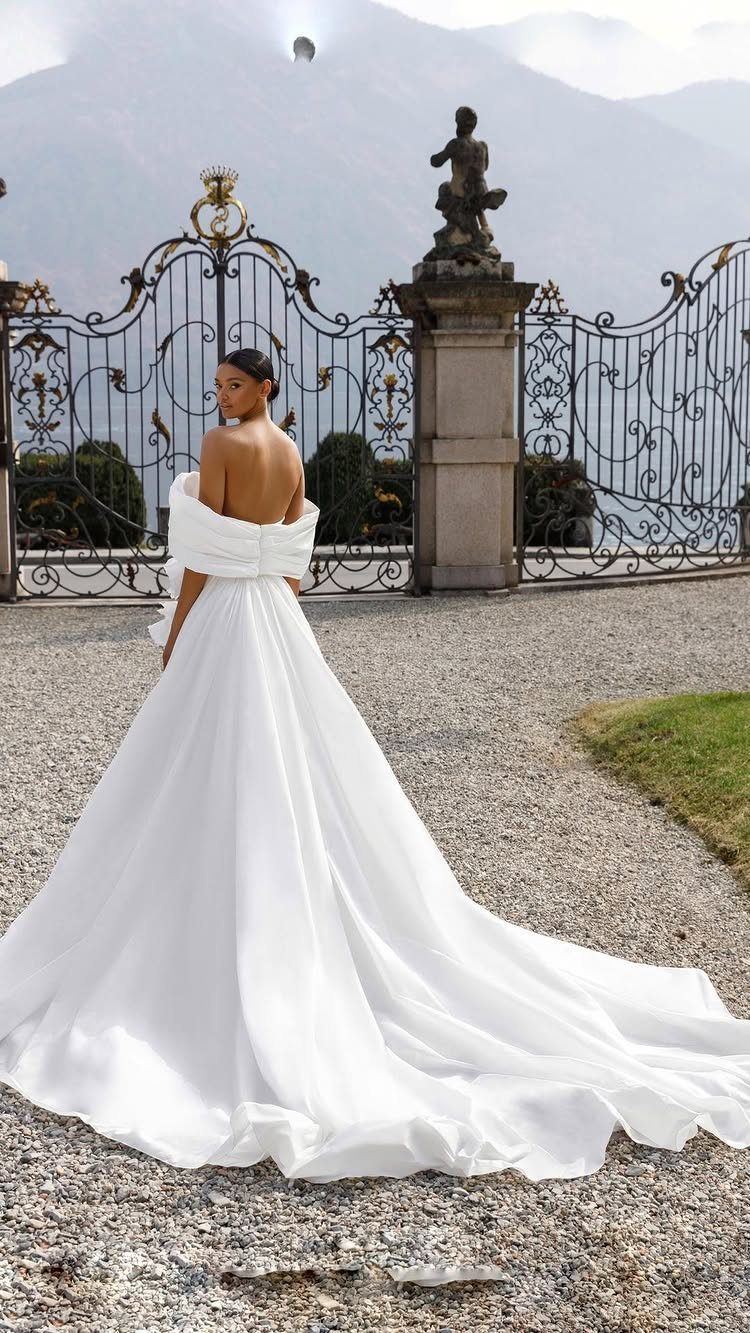 Woman in a voluminous white off-the-shoulder wedding gown standing before ornate iron gates.