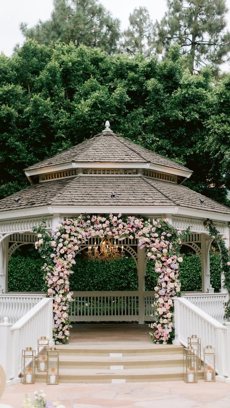White wooden gazebo adorned with pink and white floral arches and a gold chandelier.