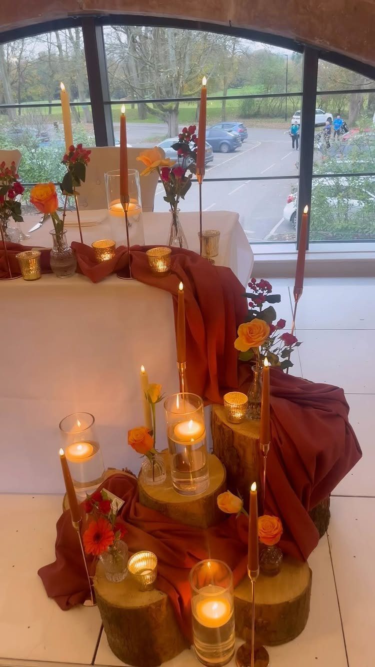 Rust-colored fabric table runner with wooden logs, orange candles, and autumn flowers.