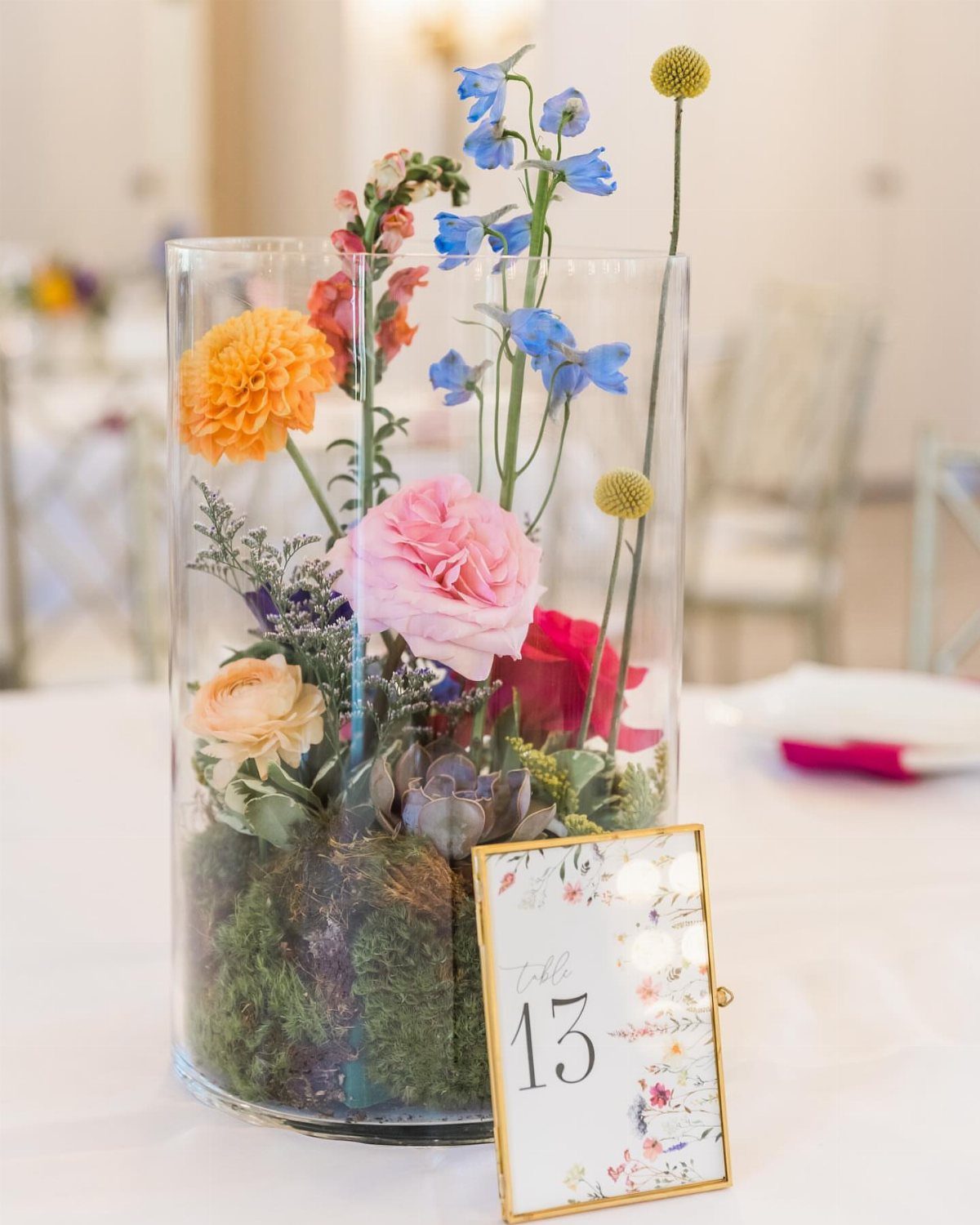 Cylindrical glass terrarium with colorful flowers and moss next to a table number card.