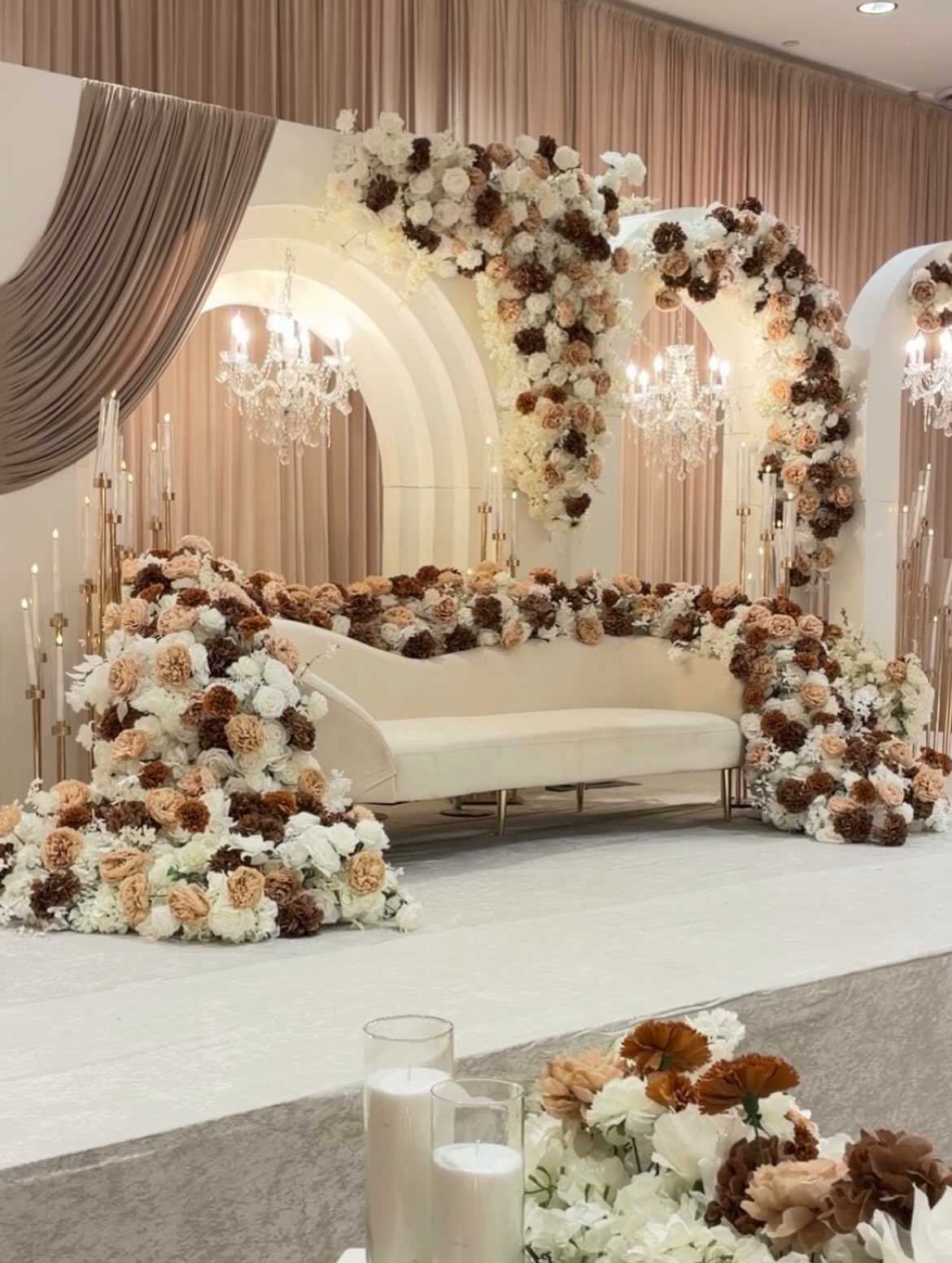 Cream curved sofa surrounded by cream and brown floral arrangements on a wedding stage