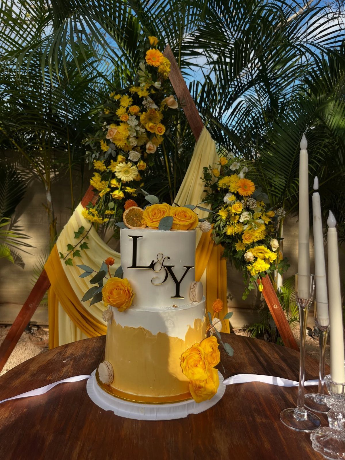 Yellow wedding cake with roses on a wooden table before a floral arch.