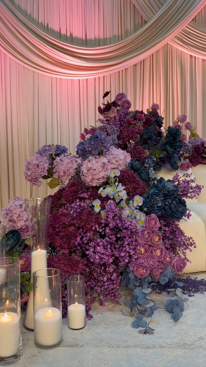 Purple and blue floral arrangement with white candles against a draped fabric backdrop.