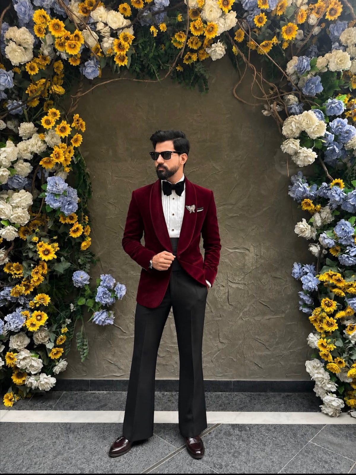 Man in a red velvet tuxedo jacket and black trousers posing by floral arch.