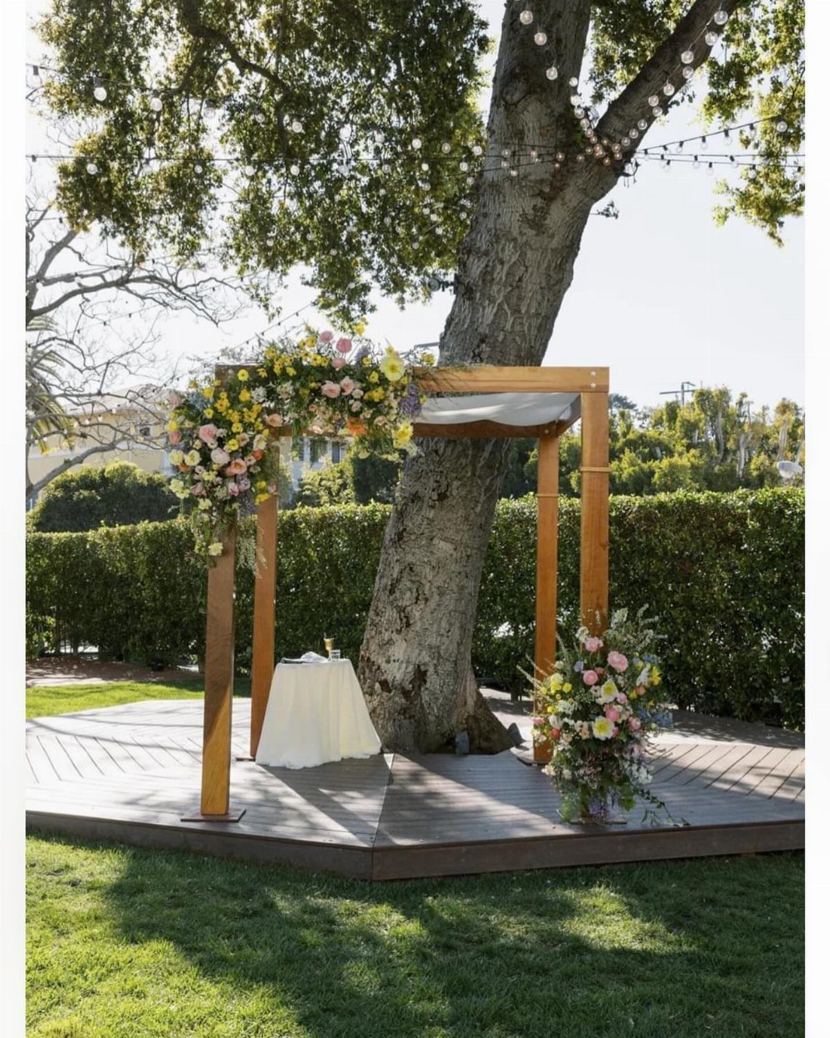 Wooden wedding chuppah with floral arrangements standing on a platform under a tree.