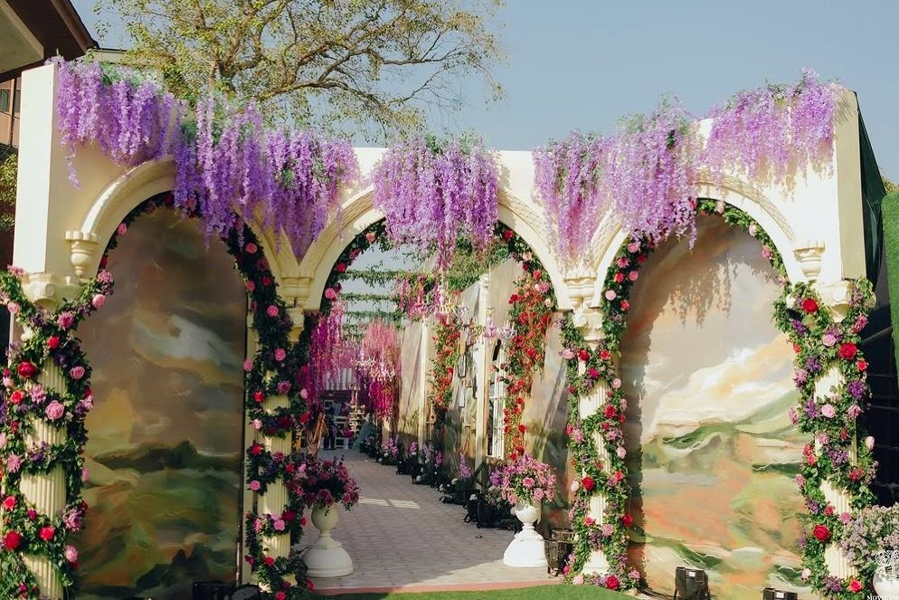 Cream architectural arches adorned with cascading purple wisteria and climbing red roses.